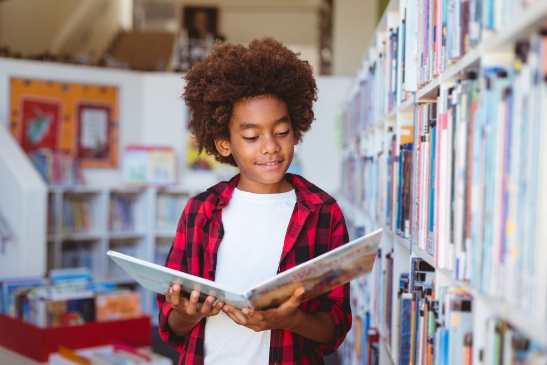 Smiling african american schoolboy reading book standing in school library